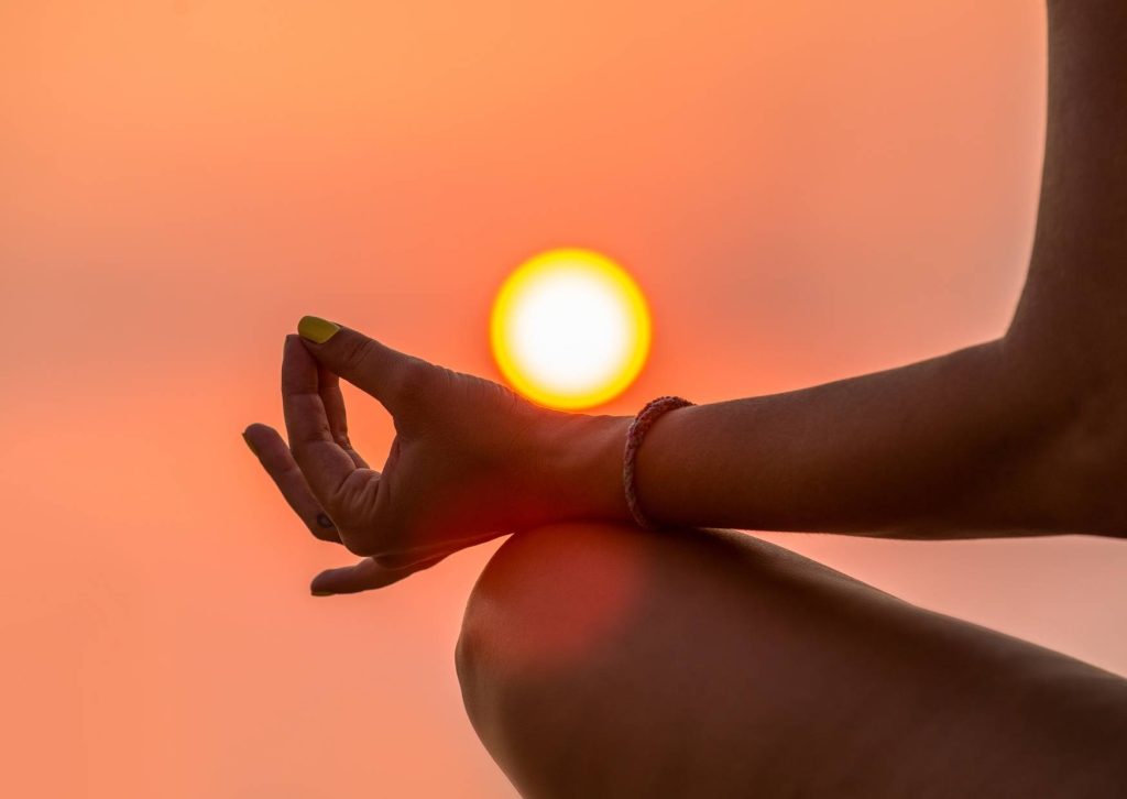 close-up of a person meditating at sunrise in Bali with hand in mudra and the sun glowing in the background on the blog how to meditate