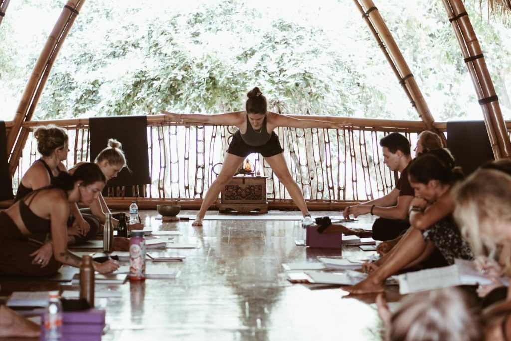 Yoga teacher training class in a bamboo shala in Bali with students taking notes during an alignment lesson