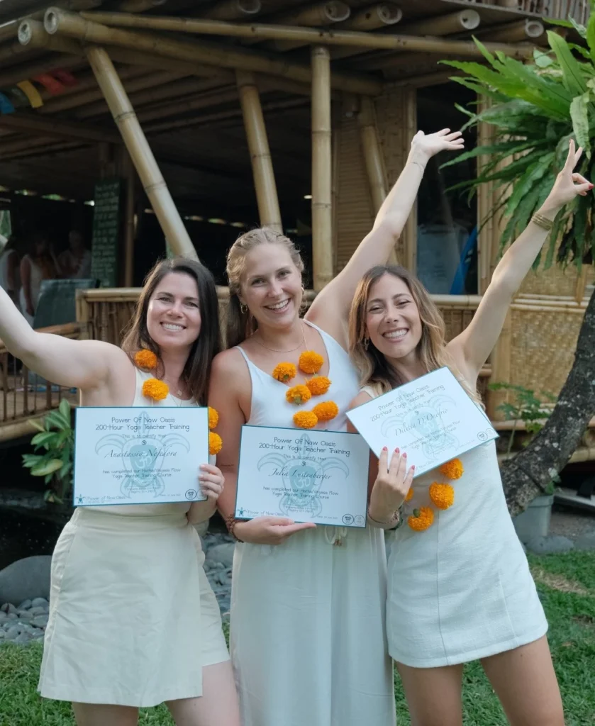 Three Yoga Teacher Training graduates celebrating with certificates and flower garlands at Power of Now Oasis in Bali