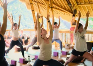 Group of Yoga Teacher Training students practicing Warrior I pose with arms raised in the bamboo shala at Power of Now Oasis in Sanur, Bali