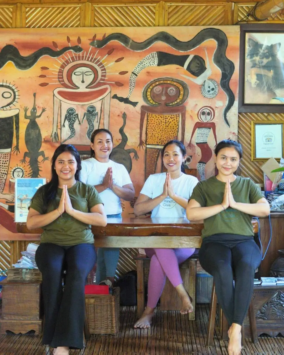 Four women from the Power of Now Oasis team sitting at the front desk in the bamboo shala, smiling and holding the namaste gesture in front of a colorful Balinese mural in Sanur, Bali