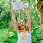 Happy Yoga Teacher Training graduate at Power of Now Oasis in Bali holding certificate and celebrating in tropical garden
