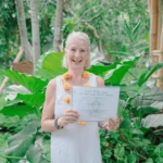 Yoga Teacher Training graduate at Power of Now Oasis in Bali smiling and holding certificate in tropical garden