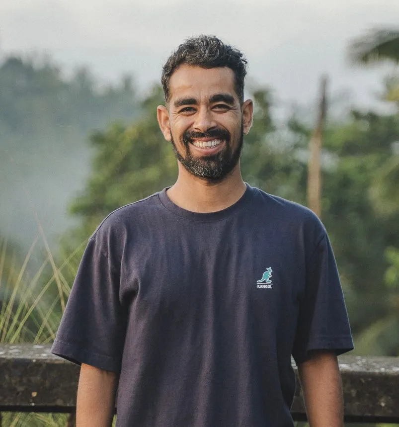 Chetan, yoga teacher at Power of Now Oasis, smiling outdoors in a navy blue shirt with a forest background