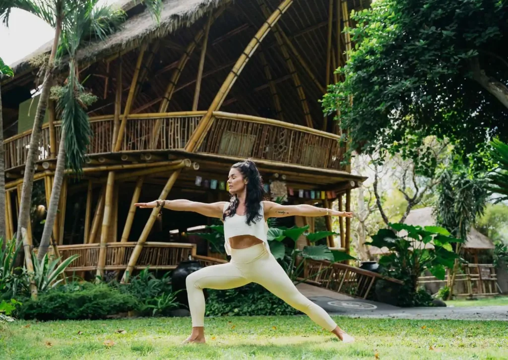 Yoga teacher practicing Warrior II pose on the lawn in front of the bamboo yoga shala at Power of Now Oasis in Sanur, Bali