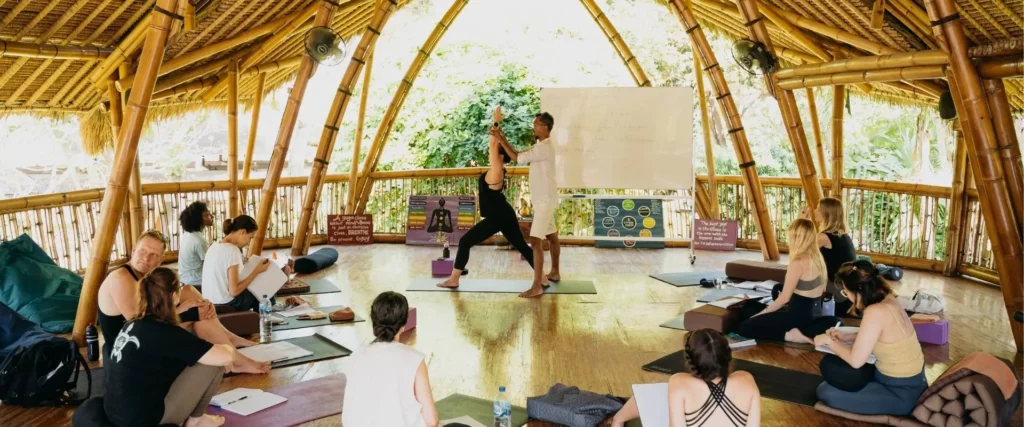 A yoga teacher adjusts a student in Warrior I pose while others observe and take notes during asana alignment class in the bamboo shala