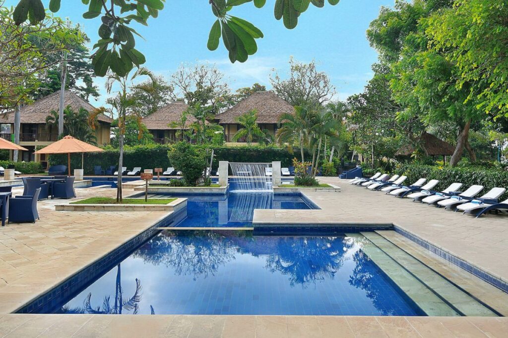 Blue-tiled pool with waterfall feature, surrounded by sun loungers and lush tropical gardens at Mercure Sanur