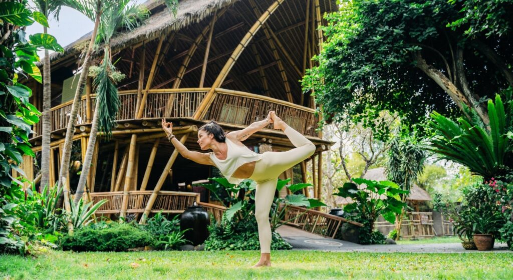 Yoga teacher in dancer pose on the lawn in front of the iconic bamboo shala at Power of Now Oasis yoga school, Sanur Bali
